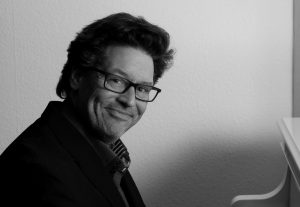 Schwarz-weißes Porträt eines Mannes mit lockigem Haar, Brille und Bart, der freundlich lächelnd über die Schulter blickt.